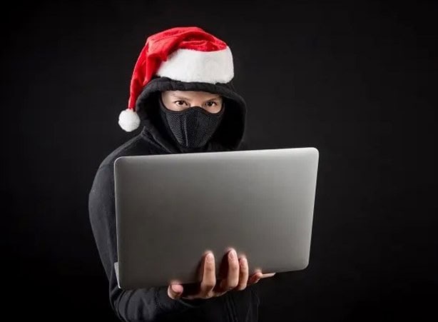 Persona con sudadera negra y mascarilla usando una laptop mientras lleva un gorro de Navidad, representando fraudes digitales en temporada decembrina.