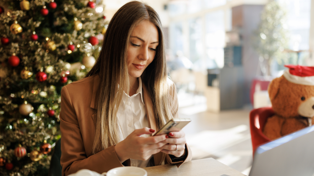 Mujer en una cafetería decorada con árbol de Navidad revisando su teléfono móvil, con un ambiente festivo y un oso de peluche con gorro navideño al fondo.